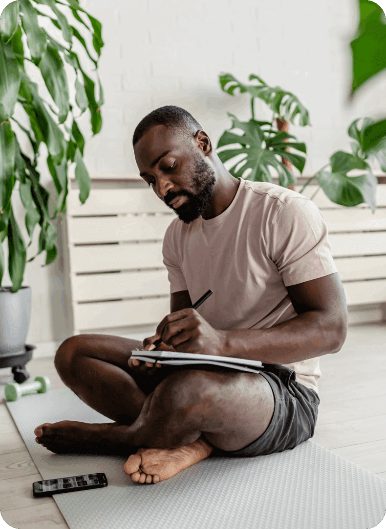 Man sitting cross-legged, writing in a notebook indoors surrounded by plants.