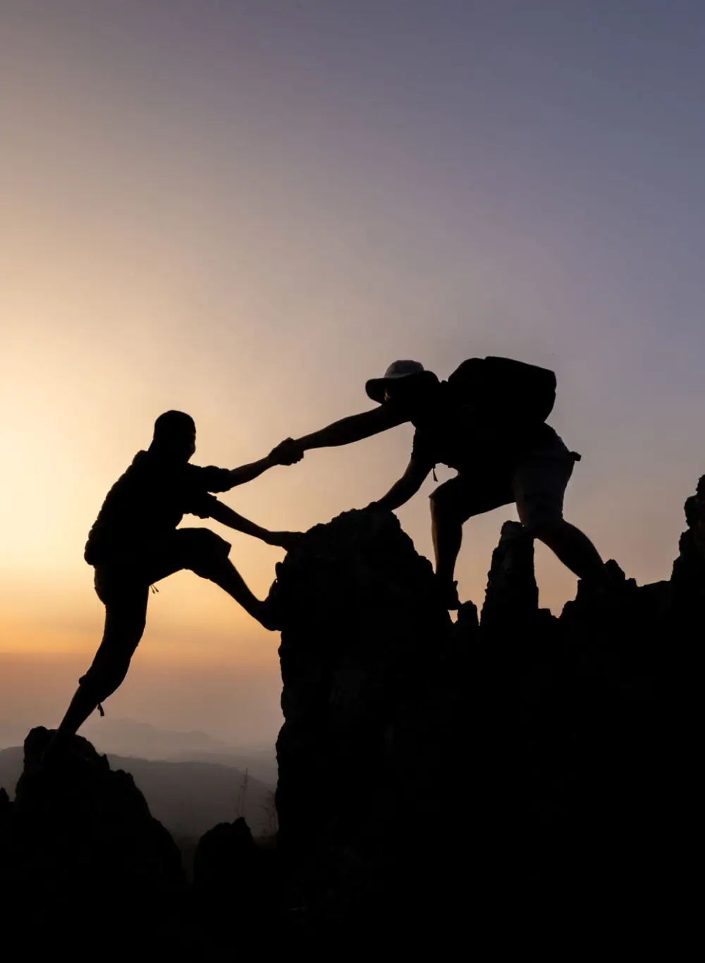 Two hikers helping each other climb a rocky peak at sunset.