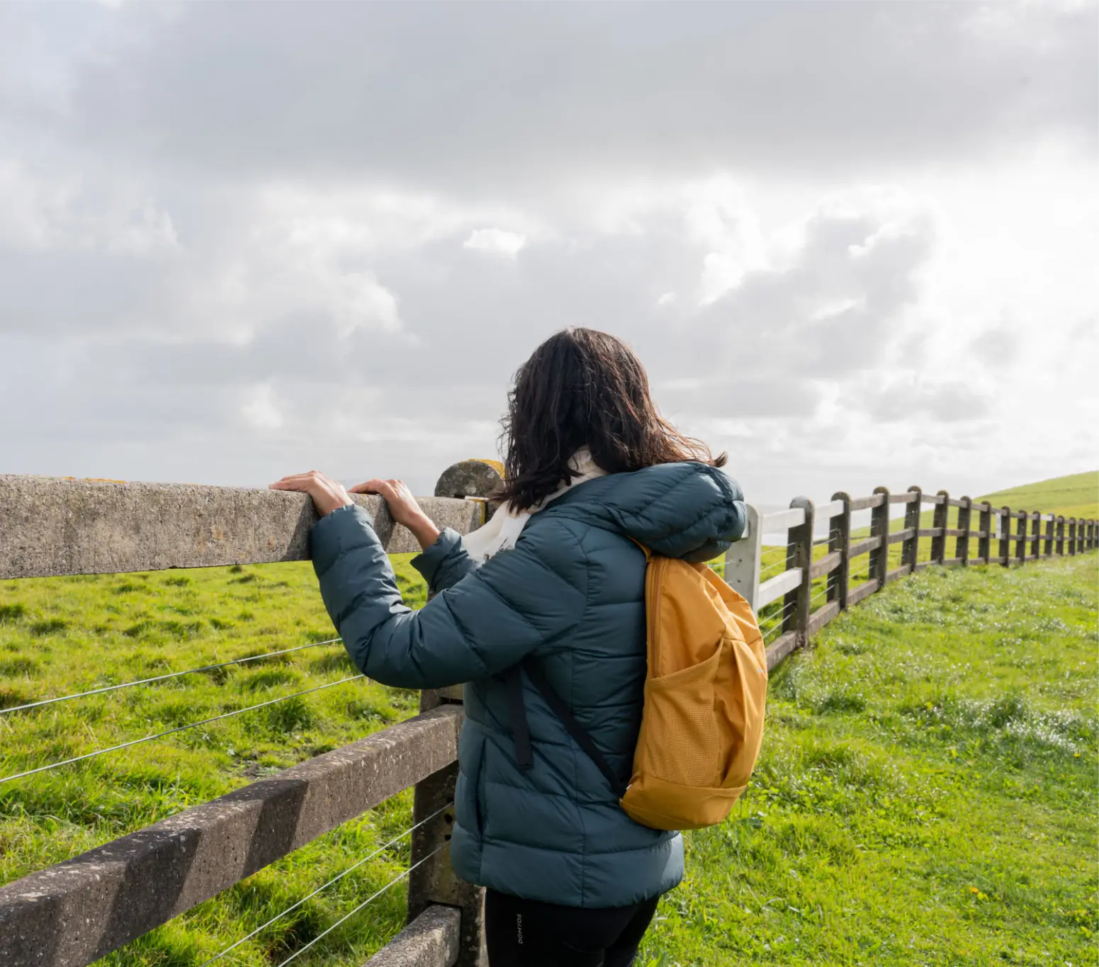 Person wearing a jacket and backpack near a wooden fence in a green field.
